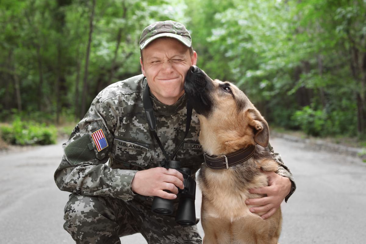 A service member in camouflage kneeling beside his dog